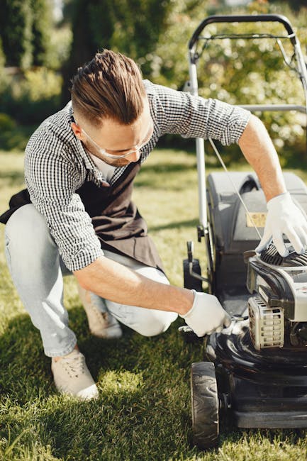 A young man wearing a checkered shirt, safety goggles, and white gloves is kneeling on a well-maintained lawn, carefully inspecting and performing garden maintenance on a lawnmower. The garden feature